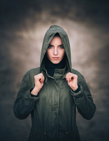 A girl in a raincoat put a hood on her head. Serious, sad, daring look. Khaki jacket on a girl.