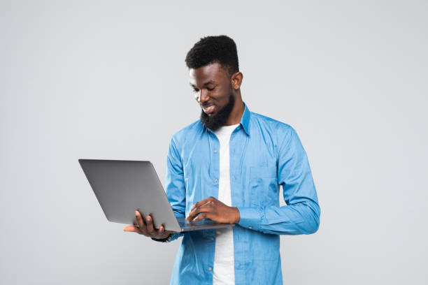 istock Young smiling african man standing and using laptop isolated over gray background 1168287267