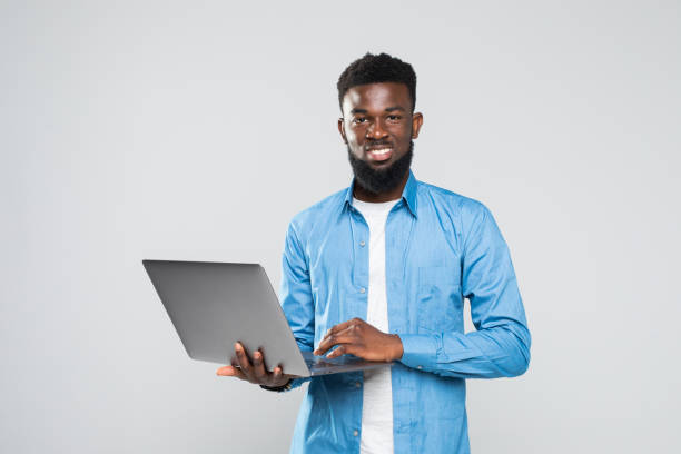 istock Young smiling african man standing and using laptop isolated over gray background 1168287276