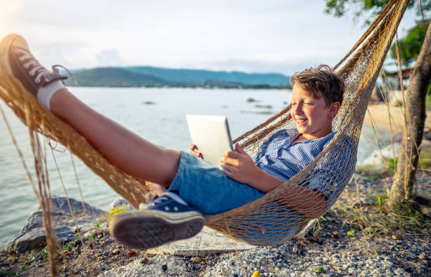 мальчик использует цифровую таблетку, лежащую в гамаке у моря - child beach digital tablet outdoors стоковые фото и изображения