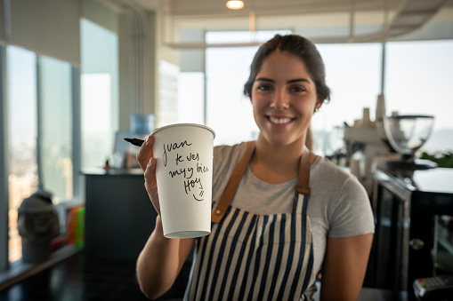Friendly beautiful woman holding a coffee cup with a personal message for customer looking at camera smiling very happy