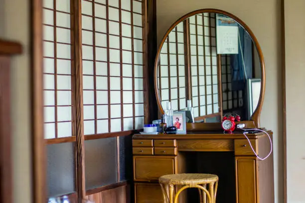 There is a woodie dresser and a rattan chair in a corner of Japanese traditional style room.  there are some of cosmetics, and a red clock on the dresser.