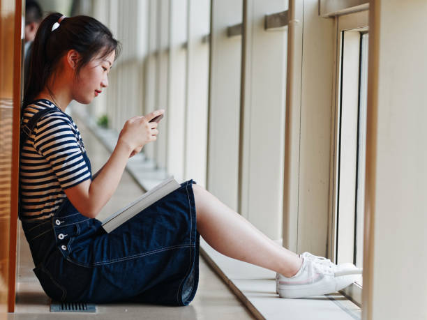 istock Young student girl with ponytail sitting on floor in library and leaning on shelf while using her smart phone with book on her legs. 1168327989