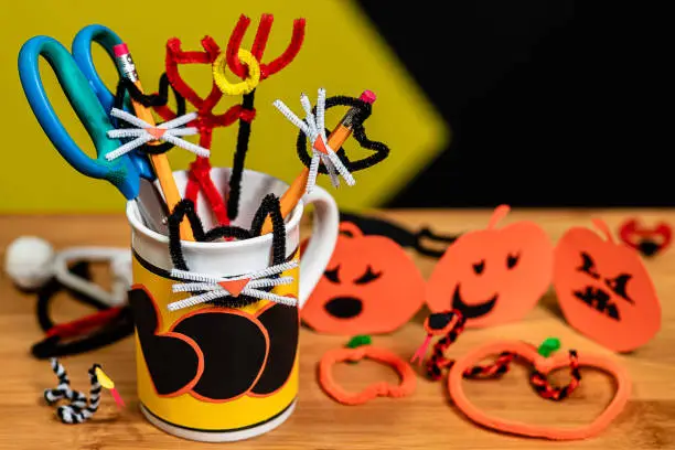 Office supplies decorated for Halloween in a Coffee Mug that says BOO. The table surface is covered in other halloween crafts.  Snakes and Jack-O-Lanterns and various other scary, spooky symbols.  negative space for Copywriting on a black field in the background.