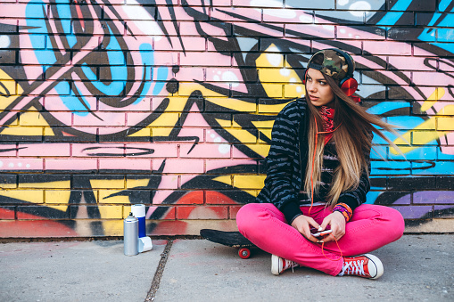 Urban young beautiful caucasian woman listening to music leaning on graffiti wall.