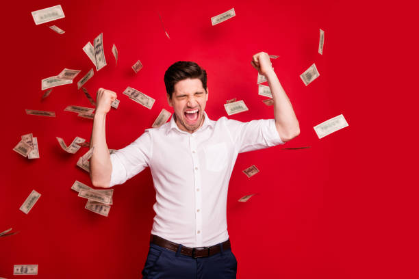 istock Photo of ecstatic overjoyed man rained with bucks banknotes achieving success while isolated with red background 1168355040