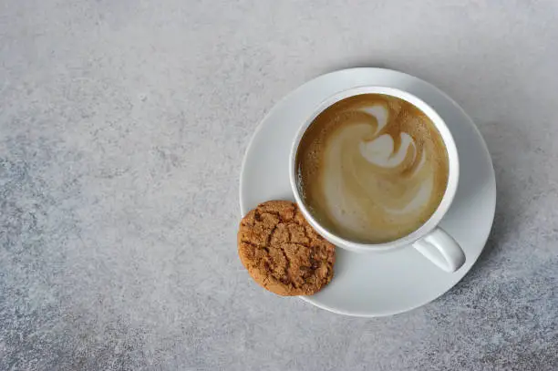 Photo of A cup of coffee on a light table surface. Next to the cup on the saucer is a cookie. Free space for text. View from above.