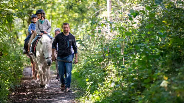 Children training horseback riding with the help of their parents in forest.