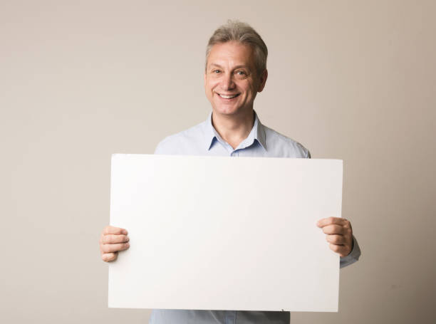 Happy mature man with blank advertising board Happy mature man with blank advertising board over grey background, free space male likeness photos stock pictures, royalty-free photos & images