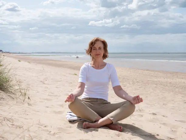 Mature woman meditating in Lotus Pose on the beach under a dramatic sky. Knokke, Belgium.