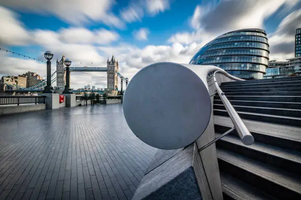 Abstract long exposure cityscape in high detail of Tower bridge crossing Thames river in London city, including City Hall at the background as photographed with extra wide lens on a sunny day with blue sky and white blurred clouds (moody sky). Shot on Canon EOS full frame system for highest quality. Long exposure technique to ensure blurred unrecognisable tourist and commuters with blurred downtown traffic.