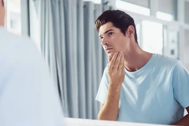 Shot of a handsome young man looking at his face in the mirror at home