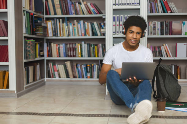 istock Afro student with headset leaning on bookshelf and using laptop 1168373159