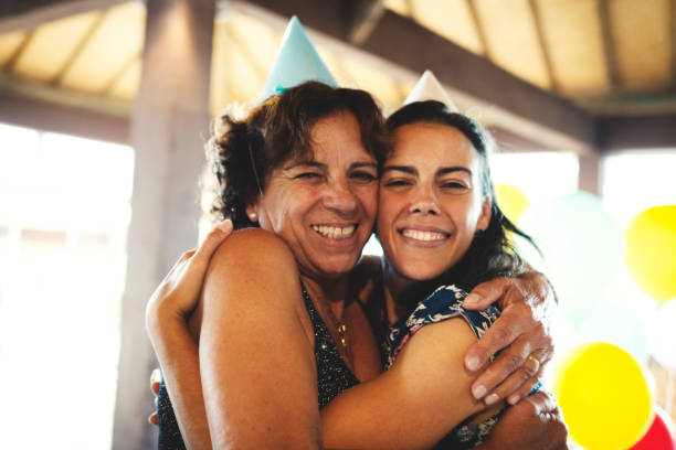 mother and daughter embracing and celebrating a birthday party using party hat - life events laughing women latin american and hispanic ethnicity imagens e fotografias de stock