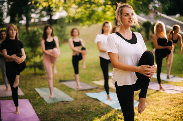 gruppe junger frauen balanciert auf einem bein und zieht ein bein hoch - yoga exercising outdoors group of people stock-fotos und bilder