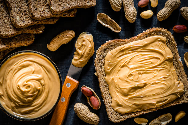 Peanut butter scattered on a slice of bread Top view of a sliced bread scattered with peanut butter on top surrounded by a kitchen knife with peanut butter on the tip, whole and peeled peanuts, some sliced breads and a bowl filled with peanut butter on a dark gray backdrop. 
Low key DSLR photo taken with Canon EOS 6D Mark II and Canon EF 24-105 mm f/4L peanutbutter stock pictures, royalty-free photos & images