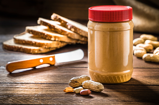 Front View of a peanut butter jar surrounded by some whole and peeled peanuts, a spreading knife and bread slices on rustic wooden table.
Low key DSLR photo taken with Canon EOS 6D Mark II and Canon EF 24-105 mm f/4L