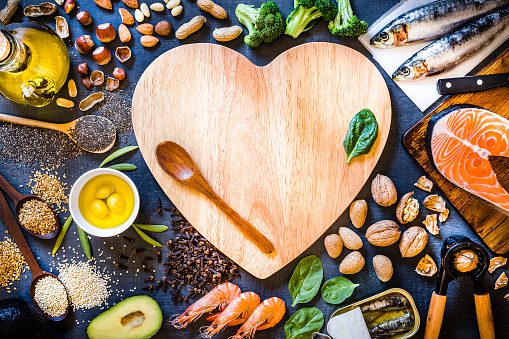 Top view of wooden cutting board with a heart shape surrounded by an assortment of food rich in Omega-3 like various kinds of nuts like hazelnuts, peanuts and almonds, canned and raw fish like salmon and sardine, some heaps of seeds like chia seeds, quinoa and flax seeds, some fruits like avocado and olives, vegetables like spinach and broccoli, and olive oil. The cutting board has a wooden spoon and a spinach leaf but also has a useful copy space on top. 
Low key DSLR photo taken with Canon EOS 6D Mark II and Canon EF 24-105 mm f/4L
