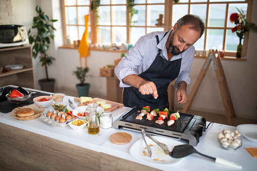 Mature man making skewers with meat and vegetables at home, on electrical barbecue