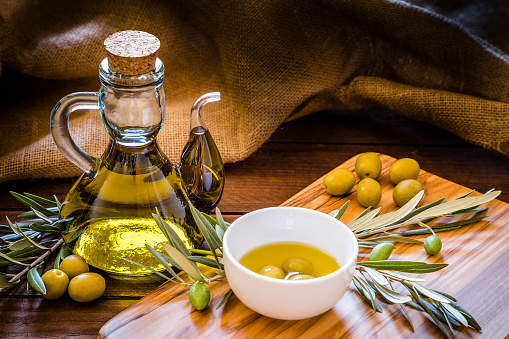 Front view of an olive oil bottle surrounded by green olives and a olive wooden cutting board with more green olives, olive branches and a withe bowl filled with three green olives and olive oil on top. Low key DSLR photo taken with Canon EOS 6D Mark II and Canon EF 24-105 mm f/4L