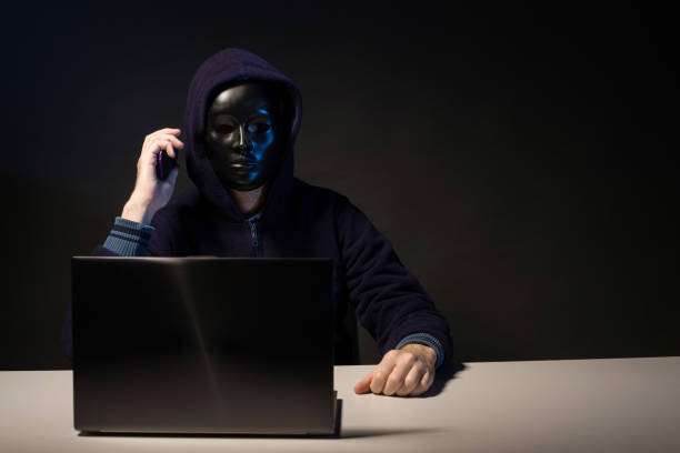 istock Anonymous hacker in mask programmer uses a laptop and talking on the phone to hack the system in the dark. 1168387401