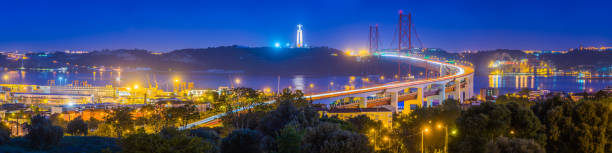 The iconic span of the 25 de Abril Bridge illuminated by the lights of the suspension cables and the zooming traffic crossing from Almada to Lisbon, Portugal’s vibrant capital city.