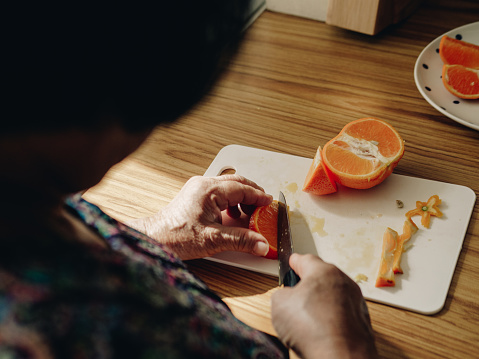 Asian Senior woman slicing fresh orange fruits on cutting board in kitchen at home.