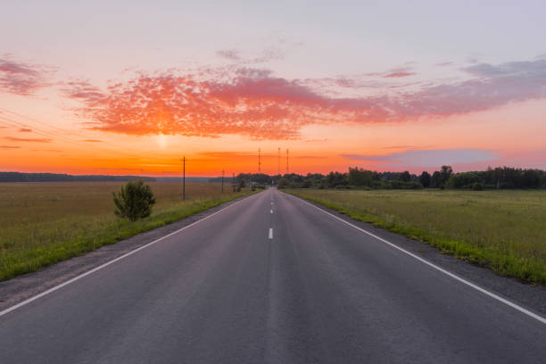 estrada asfaltada direta no meio de um campo de trigo iluminado por um nascer do sol bonito - lane sunlight sunbeam plant - fotografias e filmes do acervo