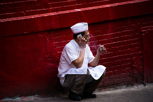 New York, United States, 2019. An asian cook sits outside and squats with cigarette while on phone.
