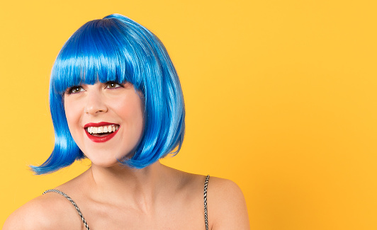 Young woman in her twenties with a great big smile.  It looks like she's looking at someone behind the photographer.  She is wearing a blue bob cut wig with a bang.    We see the straps of a summer dress.  She is smiling a big smile and is wearing red lipstick. Shot from the shoulders up.  Horizontal studio shot with a bright yellow background.  Fun, summer, funky vibe.