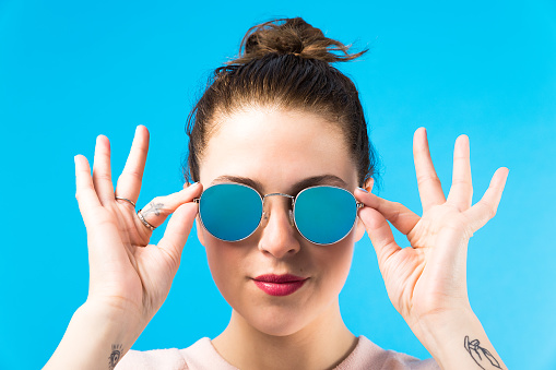 A young caucasian woman in her twenties holding mirror blue sunglasses with both hands, fingers up. She has tattoos on her index finger and both wrists and a nose hoop ring piercing. She is wearing a light pink long sleeve sweather.  Her hair is up in a bun and she is looking cool and confident. Vertical shot in studio on a blue background. It is a close-up shot from the neck up.