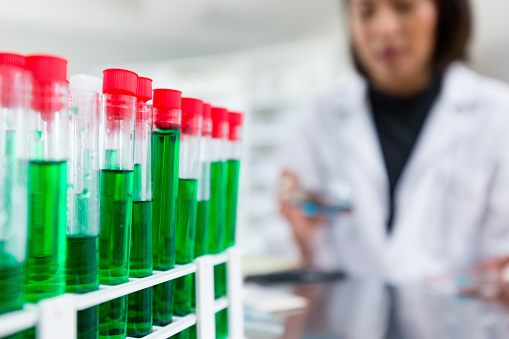Focus on test tubes in a test tube rack. A female scientist is blurred in the background.