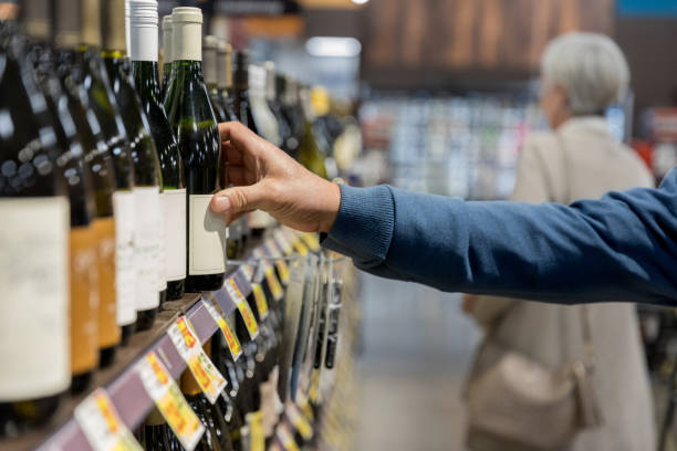 Photo of man's hand reaching for wine bottle in supermarket As his wife looks further down the aisle, the unrecognizable man reaches for a wine bottle on the shelf. alcohol shop stock pictures, royalty-free photos & images