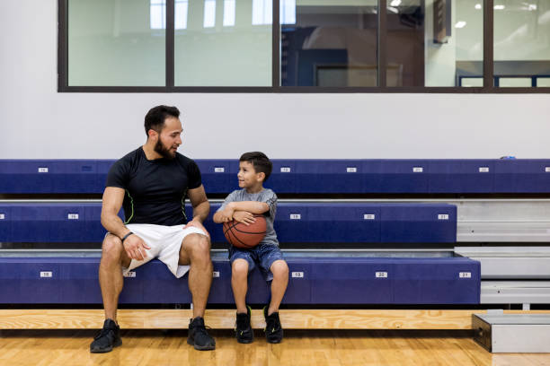 Mid adult uncle and young nephew take break at gym The mid adult uncle and his young nephew sit on the bleachers at the gym to take a break from the basketball game. school bleachers stock pictures, royalty-free photos & images