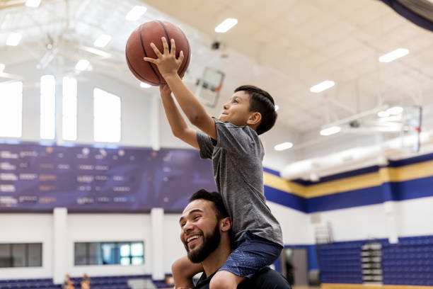 istock Sitting on dad's shoulders, young boy aims ball at basket 1168467473