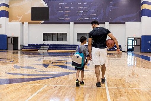 An unrecognizable father and son walk away from the basketball court in the school gym after the game.