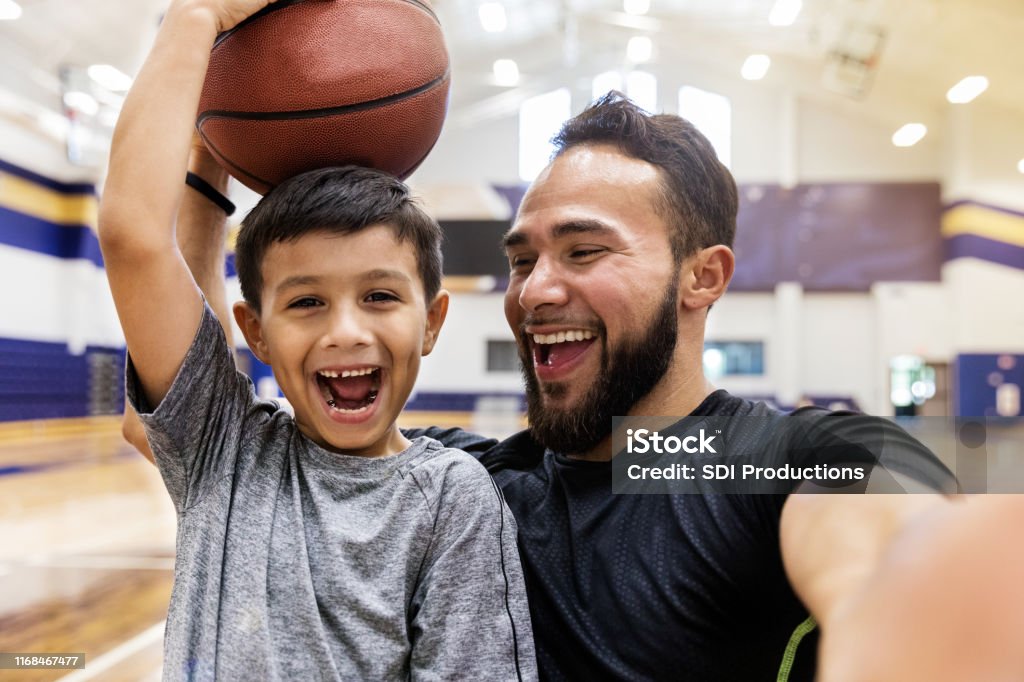 Padre se hace selfie mientras su hijo sostiene una pelota de baloncesto en la cabeza - Foto de stock de Niño libre de derechos
