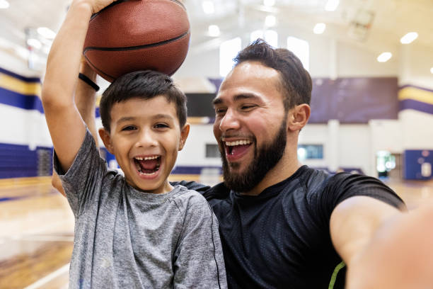 padre se hace selfie mientras su hijo sostiene una pelota de baloncesto en la cabeza - hablar fotos fotografías e imágenes de stock