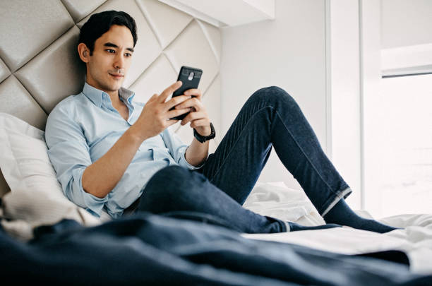 Relaxed Businessman Using Phone in Hotel Room Modern Young Taiwanese Businessman Sitting Comfortably at Hotel Room in Big Bed Using His Mobile Phone and Relaxing For a While on His Business Travel ear horn photos stock pictures, royalty-free photos & images