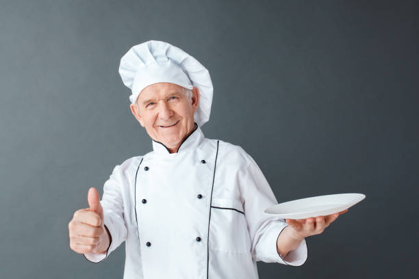 istock Senior chef studio standing isolated on gray with plate looking camera playful thumb up 1168473836