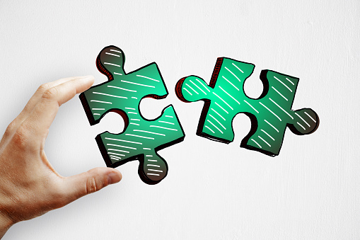 Hands holding drawn green puzzle pieces on subtle paper background. Teamwork and partnership concept