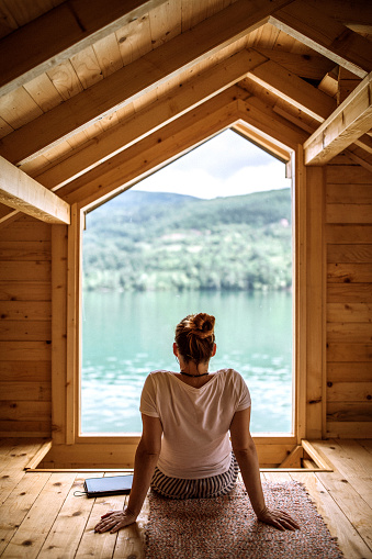 Rear view of woman sitting in wooden house on the lake and looking through window at beautiful view