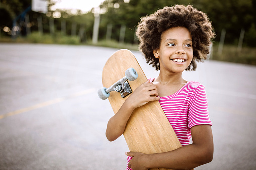 Cheerful African girl with curly hair holding skateboard and smiling happily