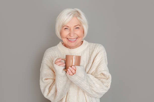 Senior woman in sweater studio isolated on grey wall drinking hot chocolate
