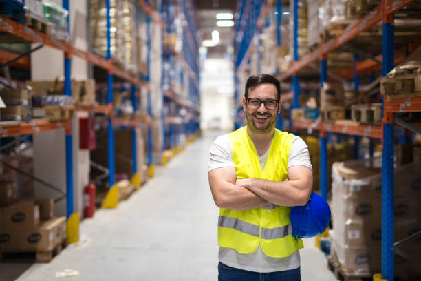 Warehouse worker. Smiling worker in distribution center. Portrait of middle aged warehouse worker standing in large warehouse distribution center with arms crossed. In background shelves with goods. reflective clothing photos stock pictures, royalty-free photos & images