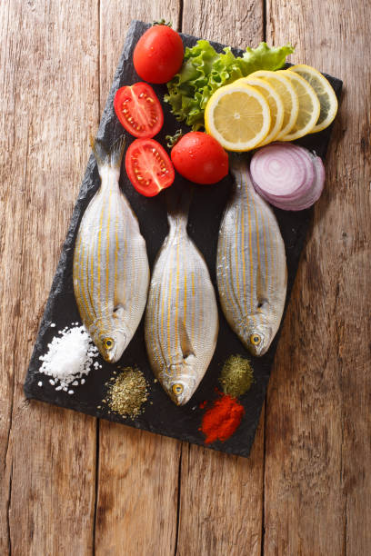 Preparation for cooking Sarpa salpa fish with lemon, vegetables and spices close-up on a slate board. Vertical top view Preparation for cooking Sarpa salpa fish with lemon, vegetables and spices close-up on a slate board on the table. Vertical top view from above salpa stock pictures, royalty-free photos & images