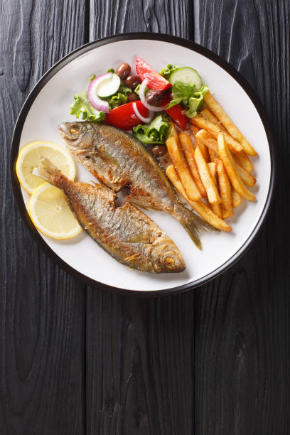 Traditional fried Sarpa salpa fish with lemon and side dish of fresh vegetable salad. french fries close-up on a plate. Vertical top view Traditional fried Sarpa salpa fish with lemon and side dish of fresh vegetable salad and french fries close-up on a plate on the table. Vertical top view from above salpa stock pictures, royalty-free photos & images