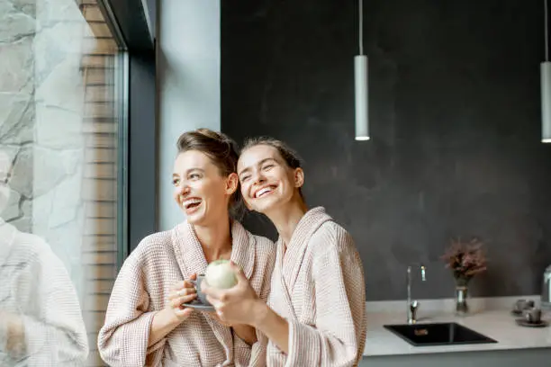 Portrait of a two young women in bathrobes standing together on the kitchen near the window, relaxing after the SPA procedures