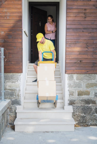 Delivery Man Pushes Dolly With Boxes To Home. A delivery man out doing his rounds, dropping off packages. Could be a full time job or just a side hustle, pushes cart on staircase. mover delivery person box hand truck stock pictures, royalty-free photos & images