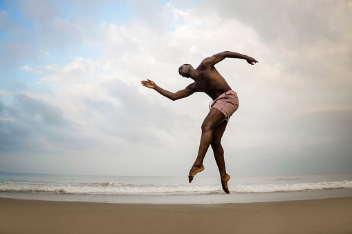 dramatic contemporary dance choreographer doing ballet beach workout . young attractive and athletic afro black American man dancing on sunrise jumping doing performance rehearsal